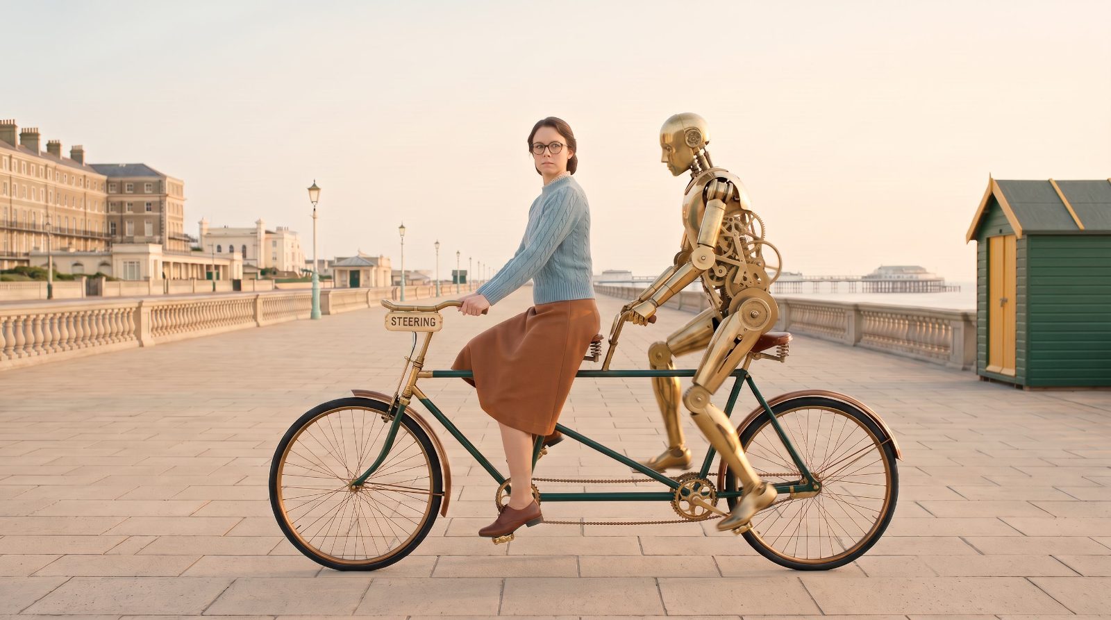 A woman riding a tandem bicycle along a seafront with a brass robot pedalling behind her